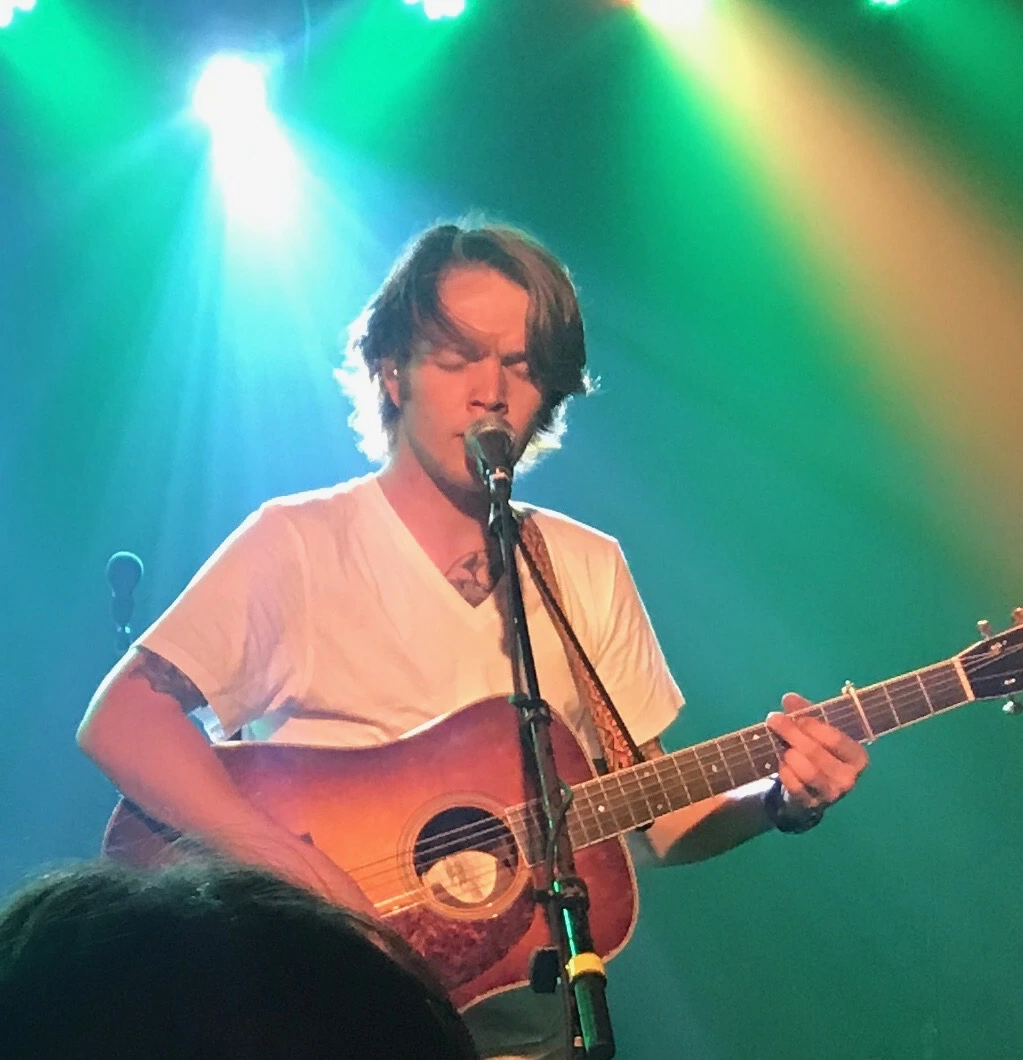 Billy Strings performing onstage with an acoustic guitar under colorful lights