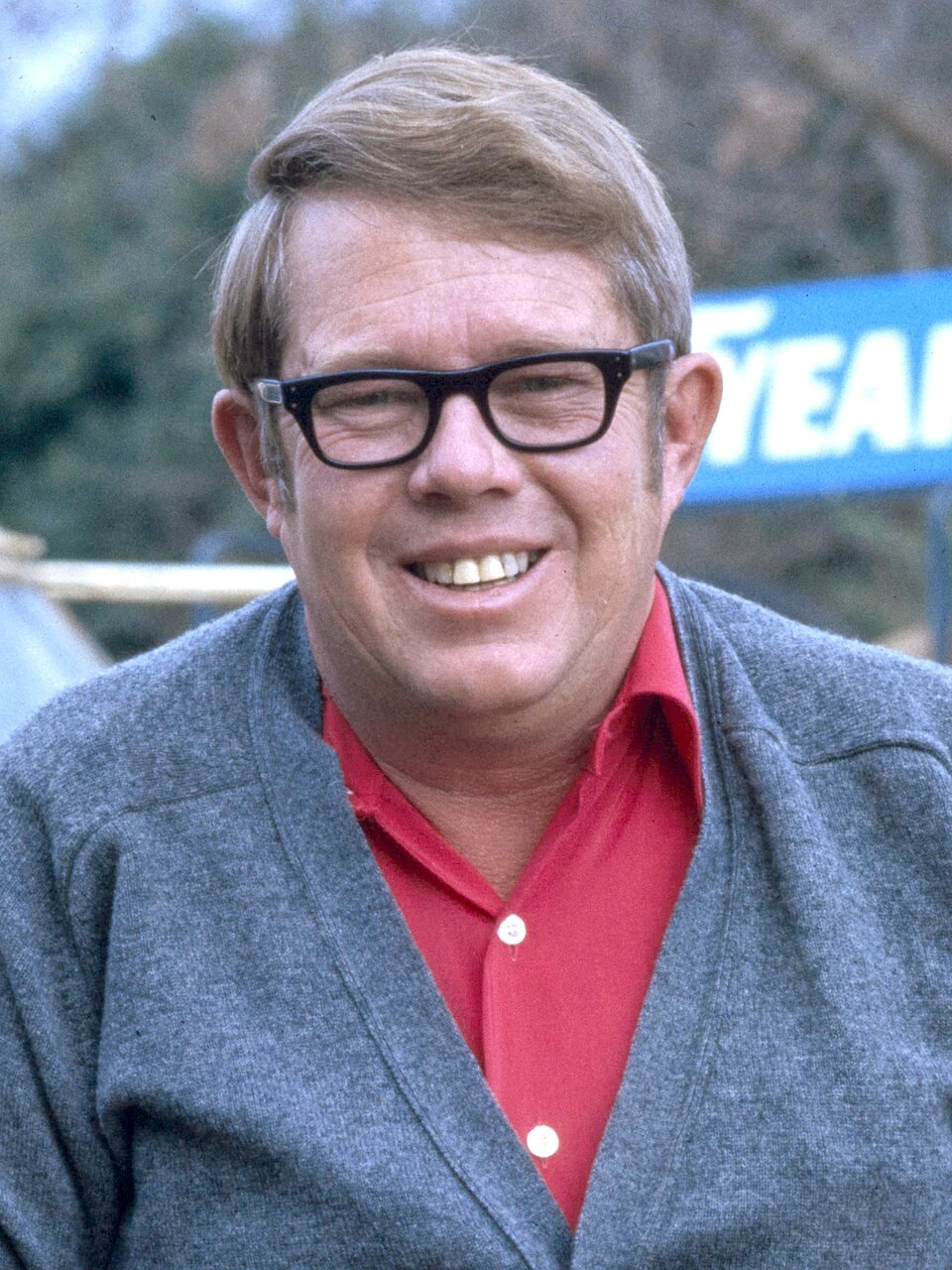 Billy Carter smiling outdoors in glasses and a gray cardigan over a red shirt.